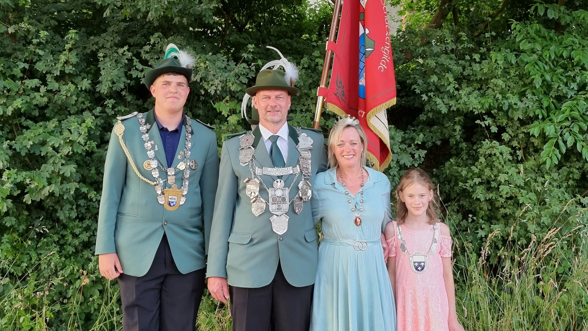 Zwei Männer, eine Frau und ein Mädchen stehen vor einer Hecke. Die Männer tragen Schützenuniform. Hinter den vier Menschen steht eine rote Fahne.