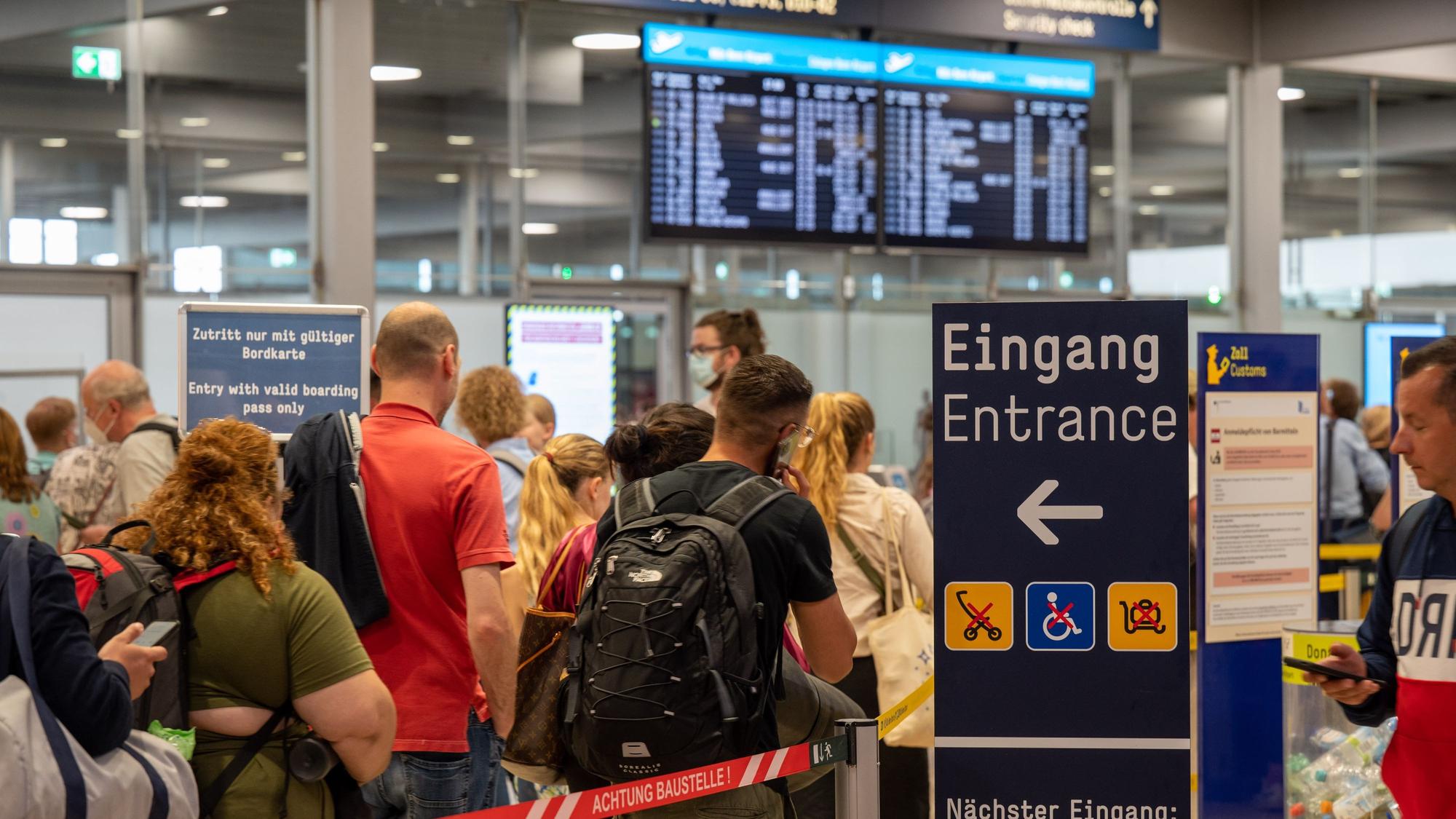 Am Flughafen Köln/Bonn warten Reisende im Sommer 2022 an der Sicherheitskontrolle. Es haben sich extrem lange Schlangen gebildet, im Hintergrund ist ein Fluganzeiger zu sehen. (Archivbild)