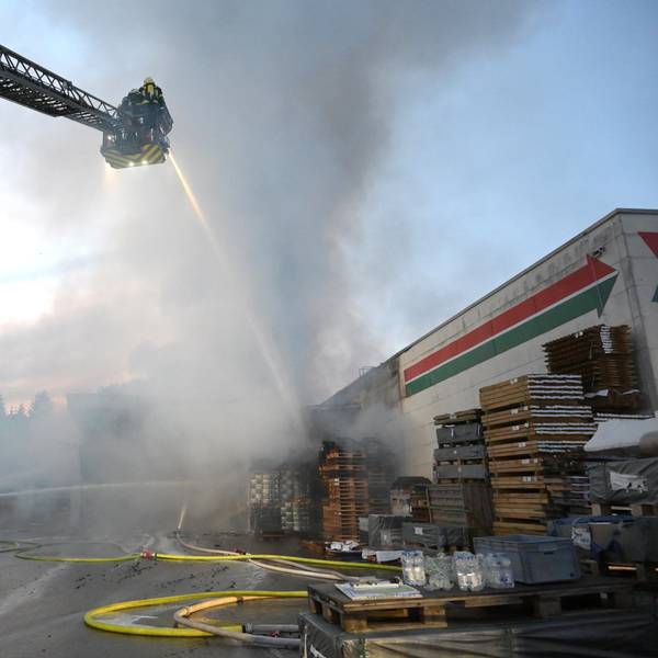 Ein Feuerwehrwagen steht vor einem brennenden Gebäude, auf der ausfahrbaren Leiter stehen Feuerwehrleute und kämpfen mit einem dicken Wasserstrahl gegen die Flammen.