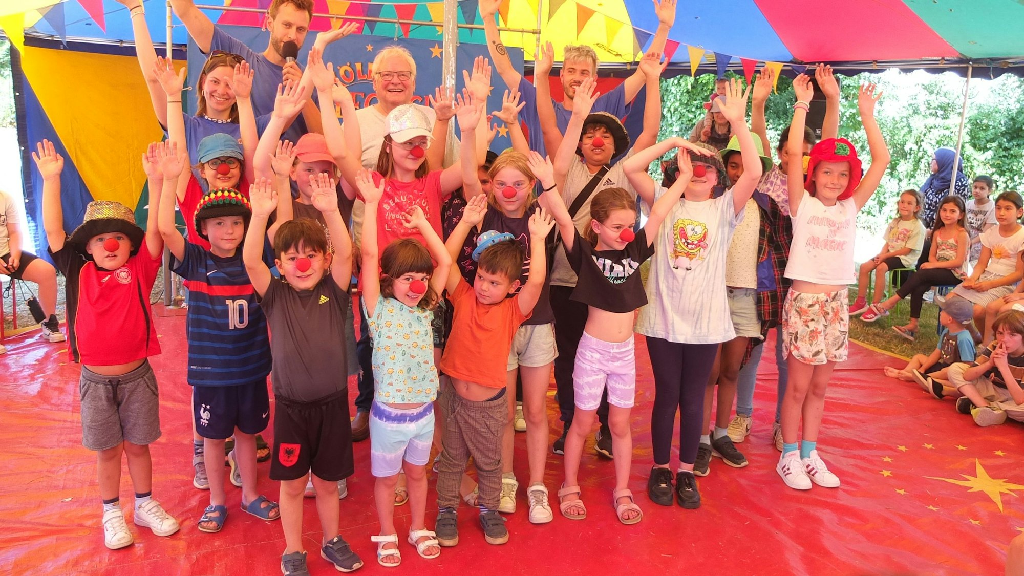 Kinder stehen winkend und mit Clownsnasen unter einem dem Dach eines bunten Zirkuszelts.