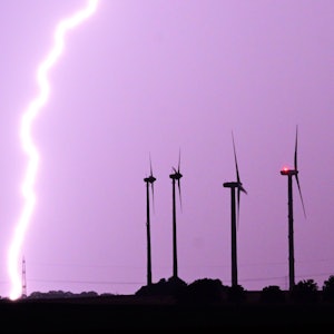 Ein Blitz entlädt sich während eines Gewitters hinter Windrädern. Der Himmel ist lila gefärbt. (Symbolbild)