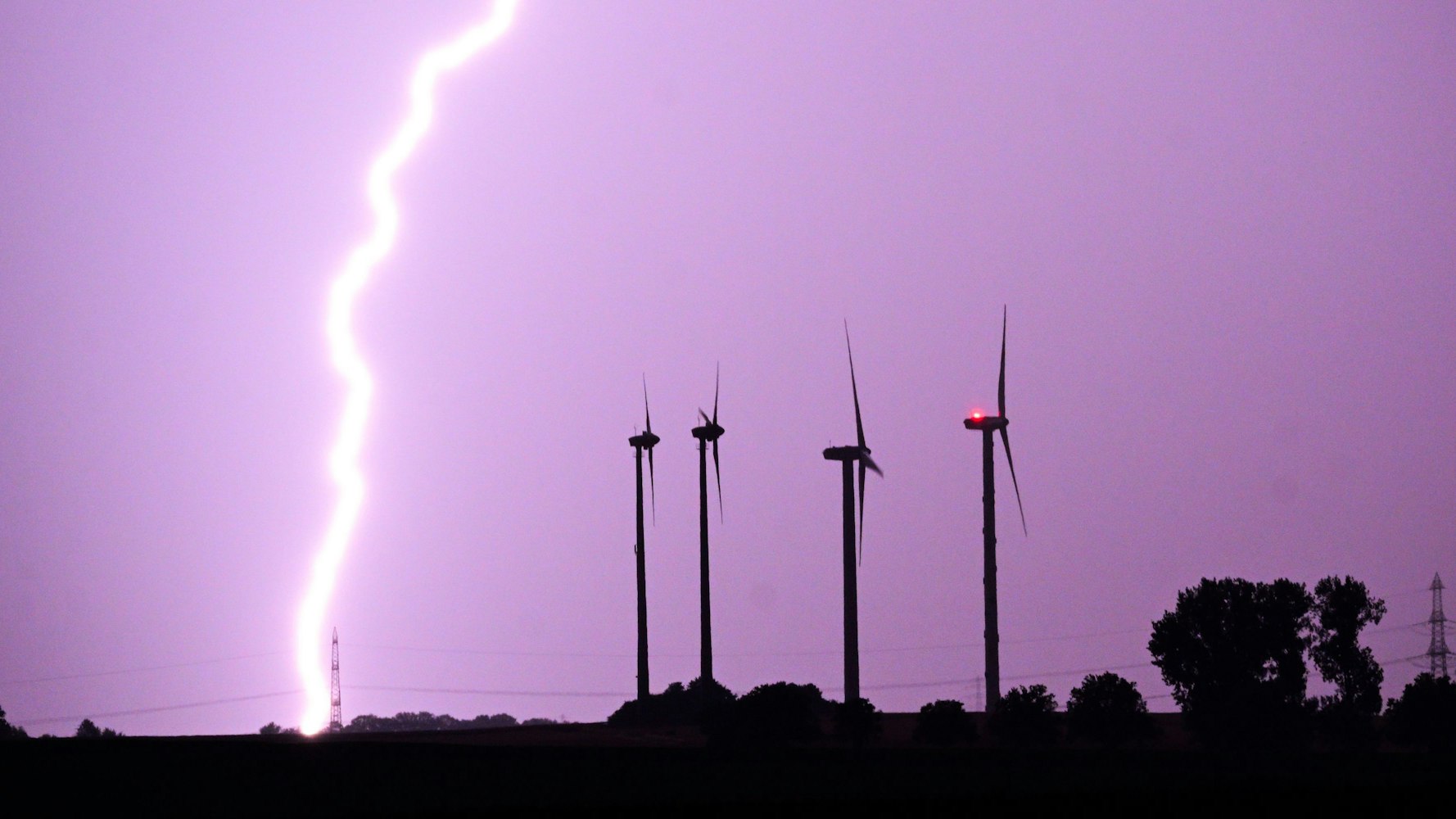 Ein Blitz entlädt sich während eines Gewitters hinter Windrädern. Der Himmel ist lila gefärbt. (Symbolbild)