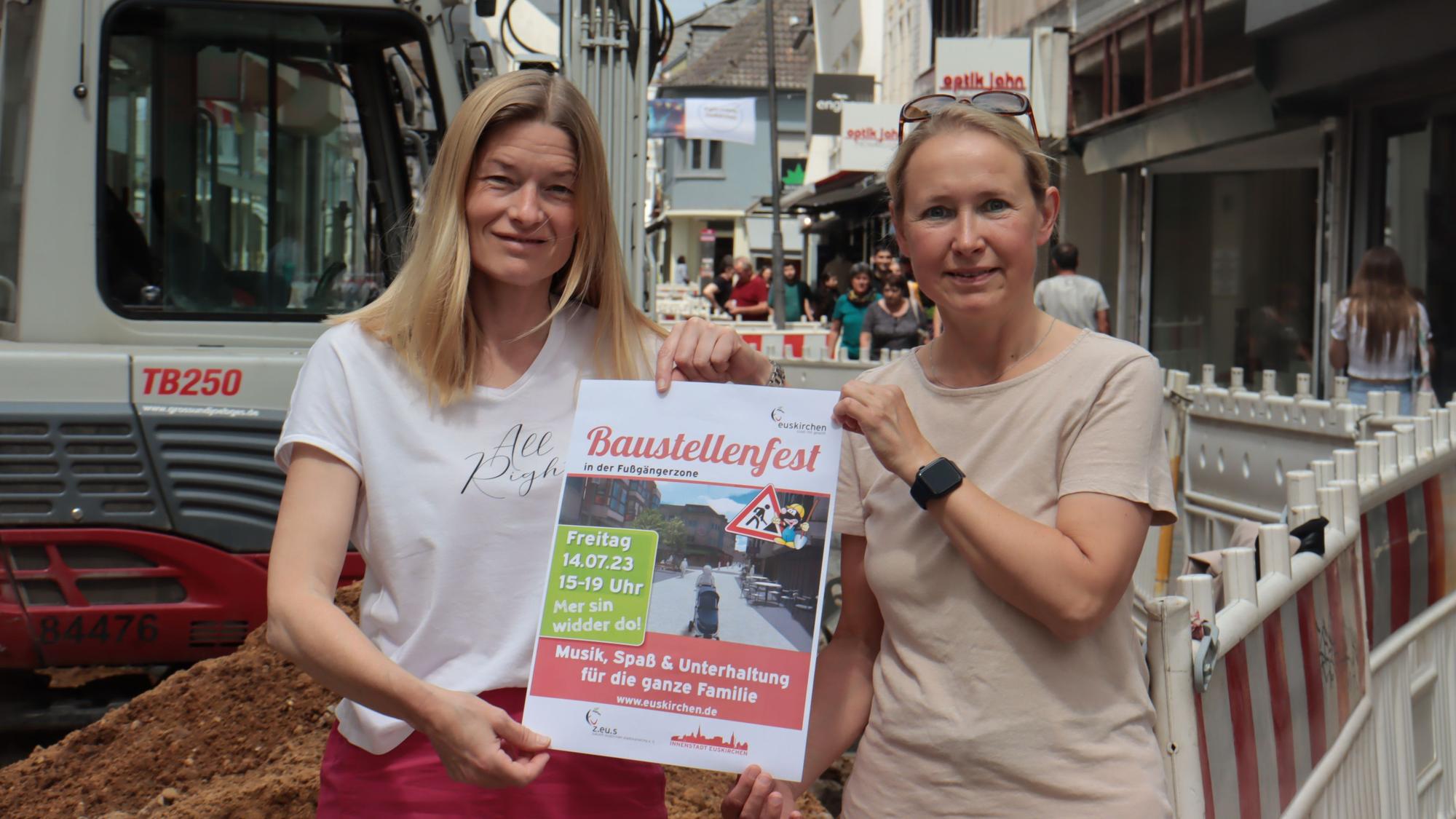 Zwei Frauen stehen vor einer Baustellenabsperrung in der Euskirchener Innenstadt und halten ein Plakat, das auf ein Fest hinweist.