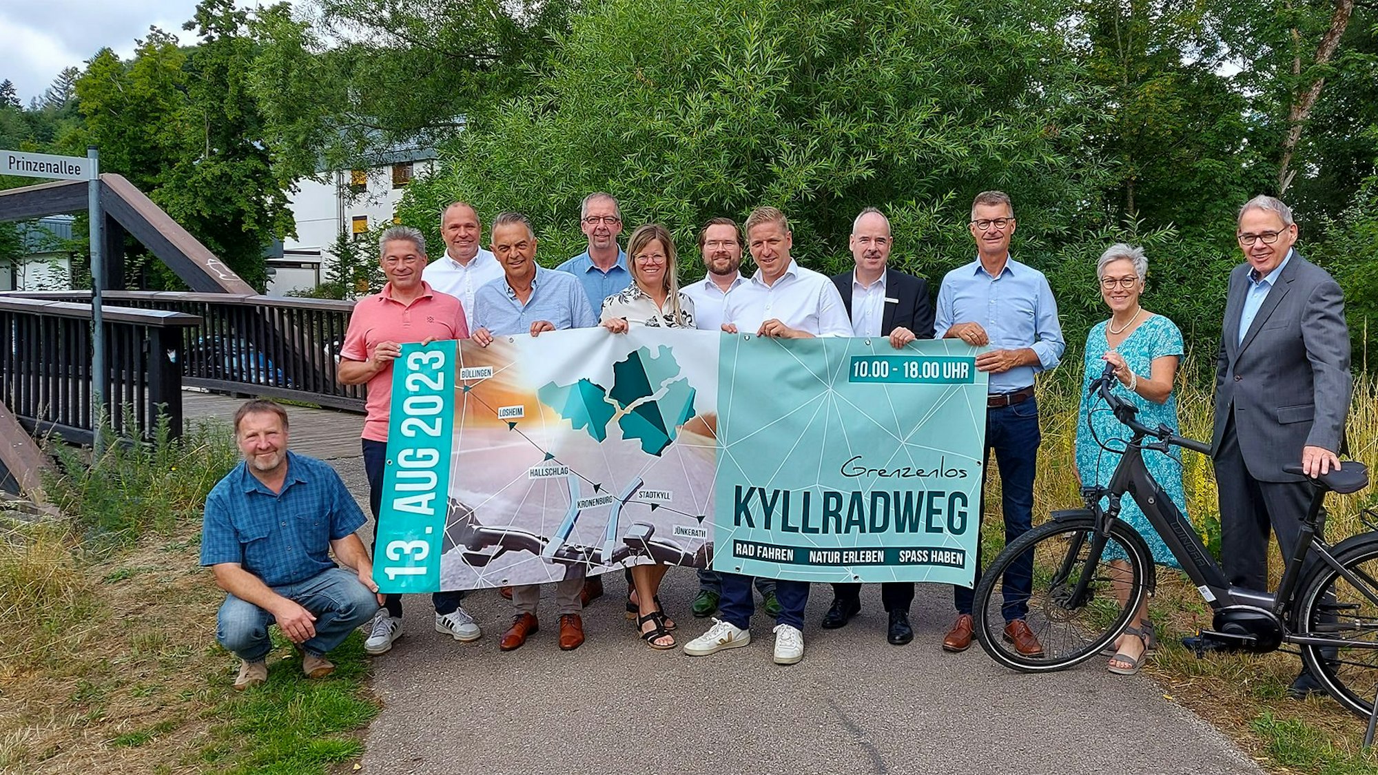 Eine Gruppe Menschen steht vor einer Brücke. Die Männer und Frauen halten ein großformatiges Plakat, mit dem auf den Radaktionstag im Kylltal hingewiesen wird.