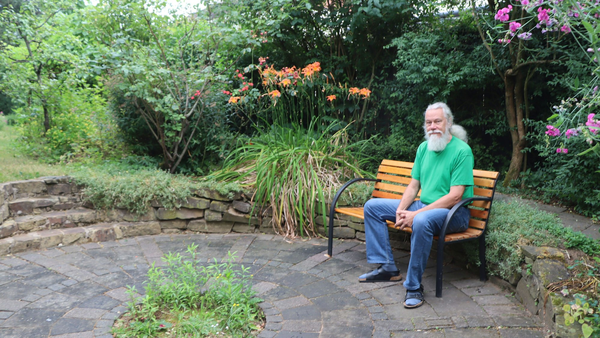 Entspannt sitzt Erich Hermans, ein Herr mit ergrautem Bart und langem Haar, auf einer Bank in seinem mit vielen Pflanzen begrünten Garten.