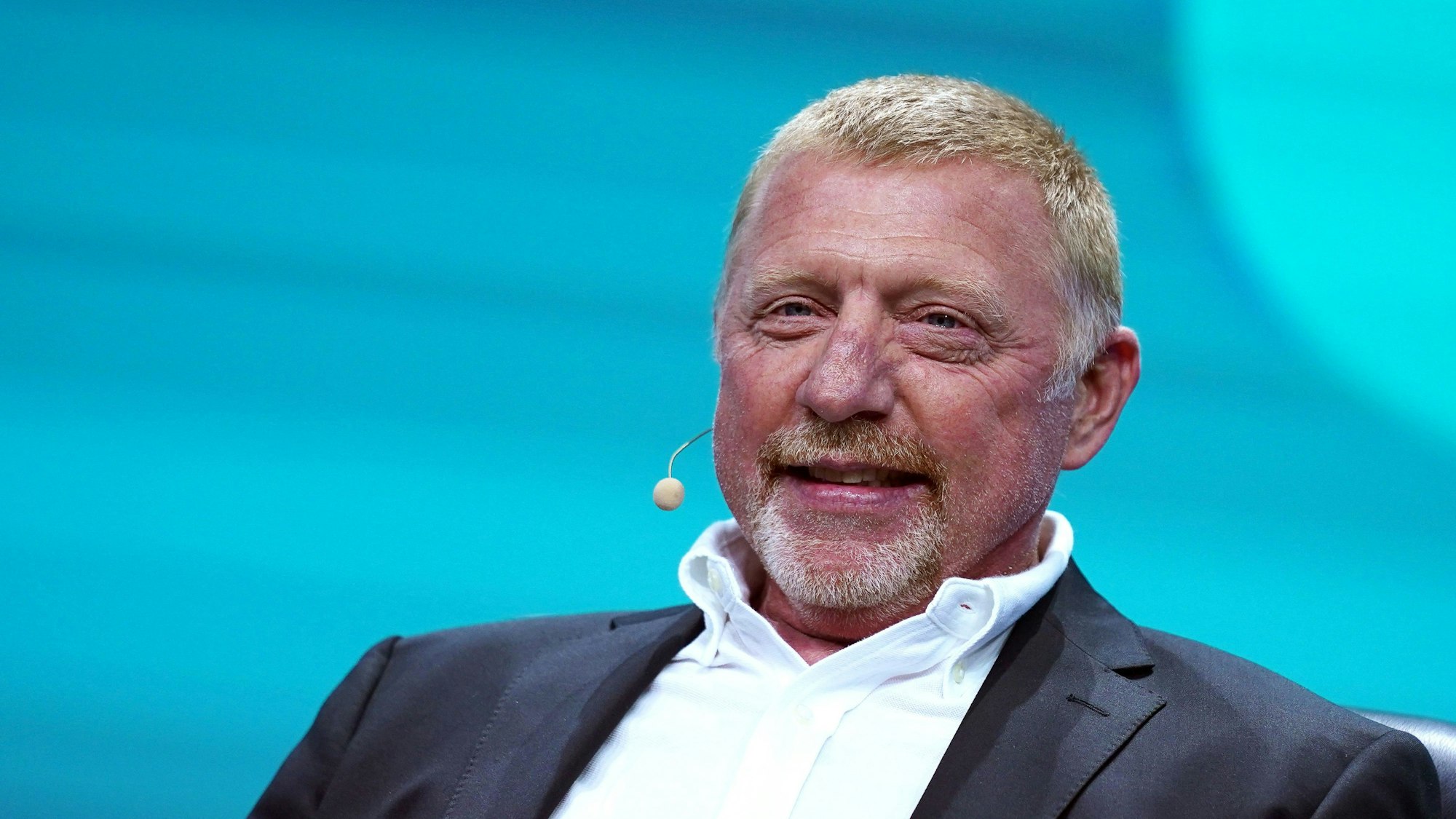 Die deutsche Tennis-Legende Boris Becker sitzt bei einer Veranstaltung auf der Bühne. Becker trägt einen Anzug und hat ein Headset aufgesetzt.