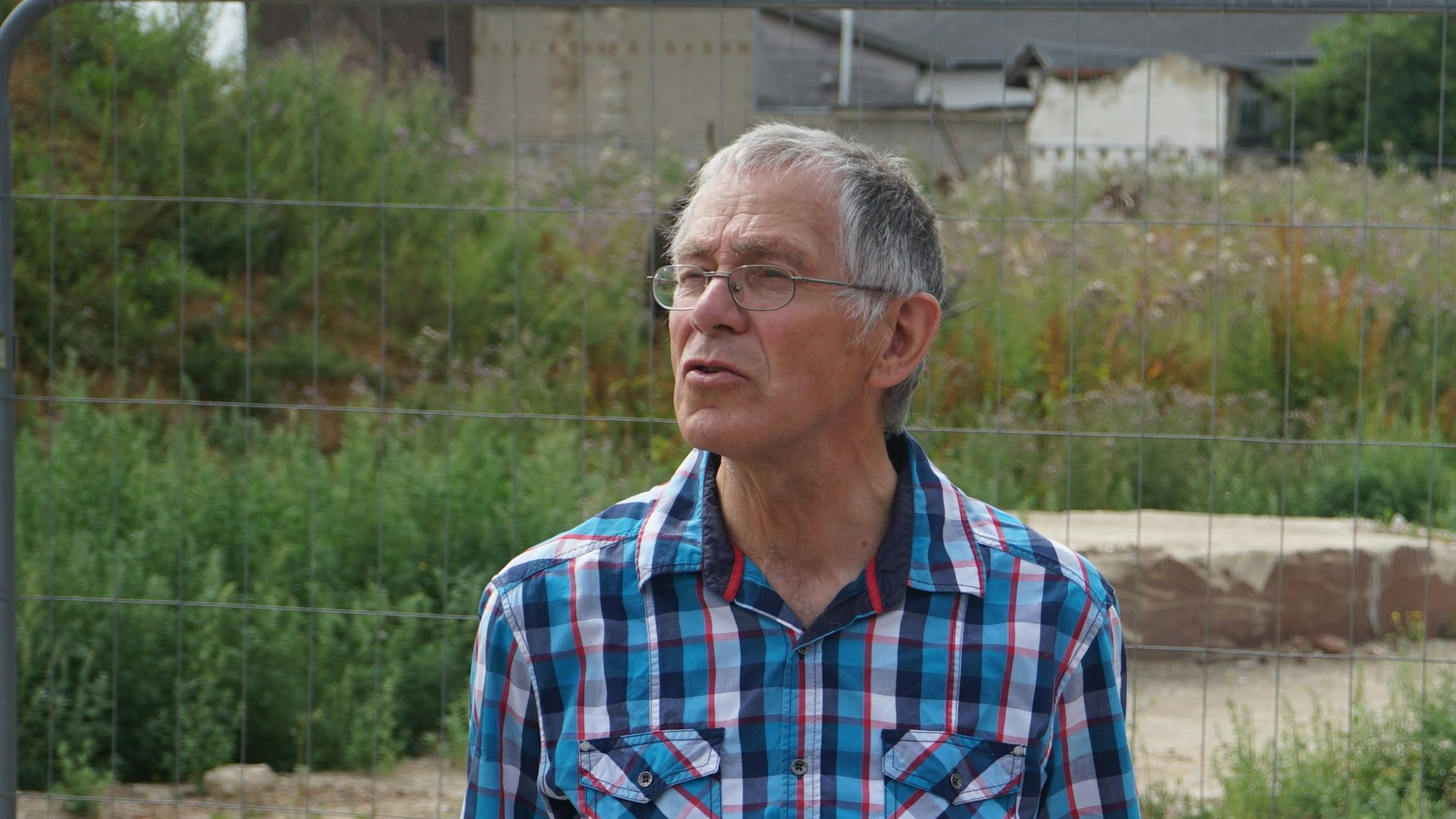 Heinrich Portz steht vor einem Bauzaun in Manheim. Dahinter sind Häuserruinen und wild wucherndes Unkraut zu sehen.