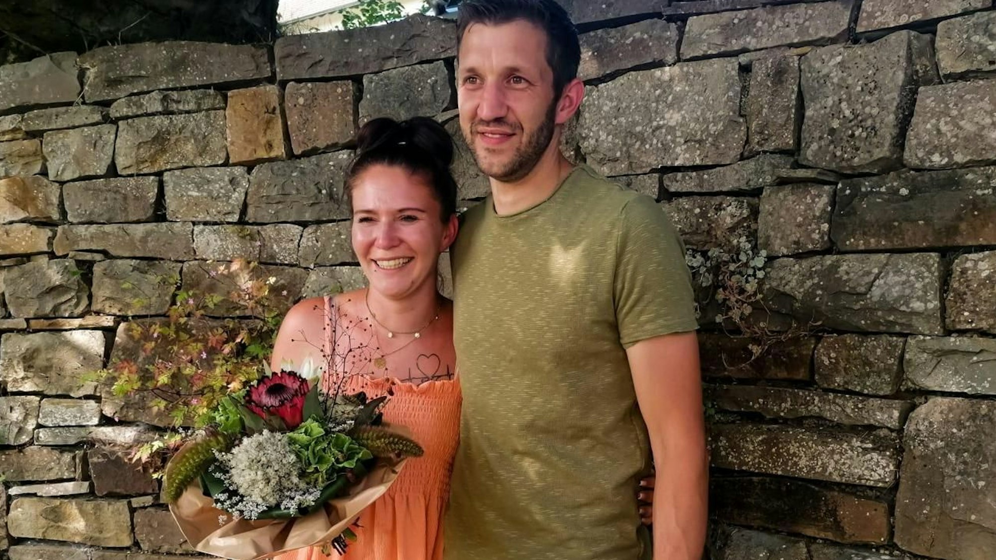 Das Bild zeigt ein junges Paar vor einer alten Mauer.