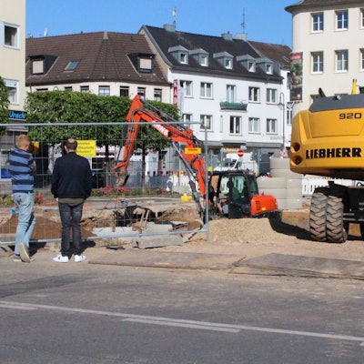 Zwei Männer schauen durch einen Bauzaum auf eine Baustelle. Mehrere Bagger sind zu sehen.