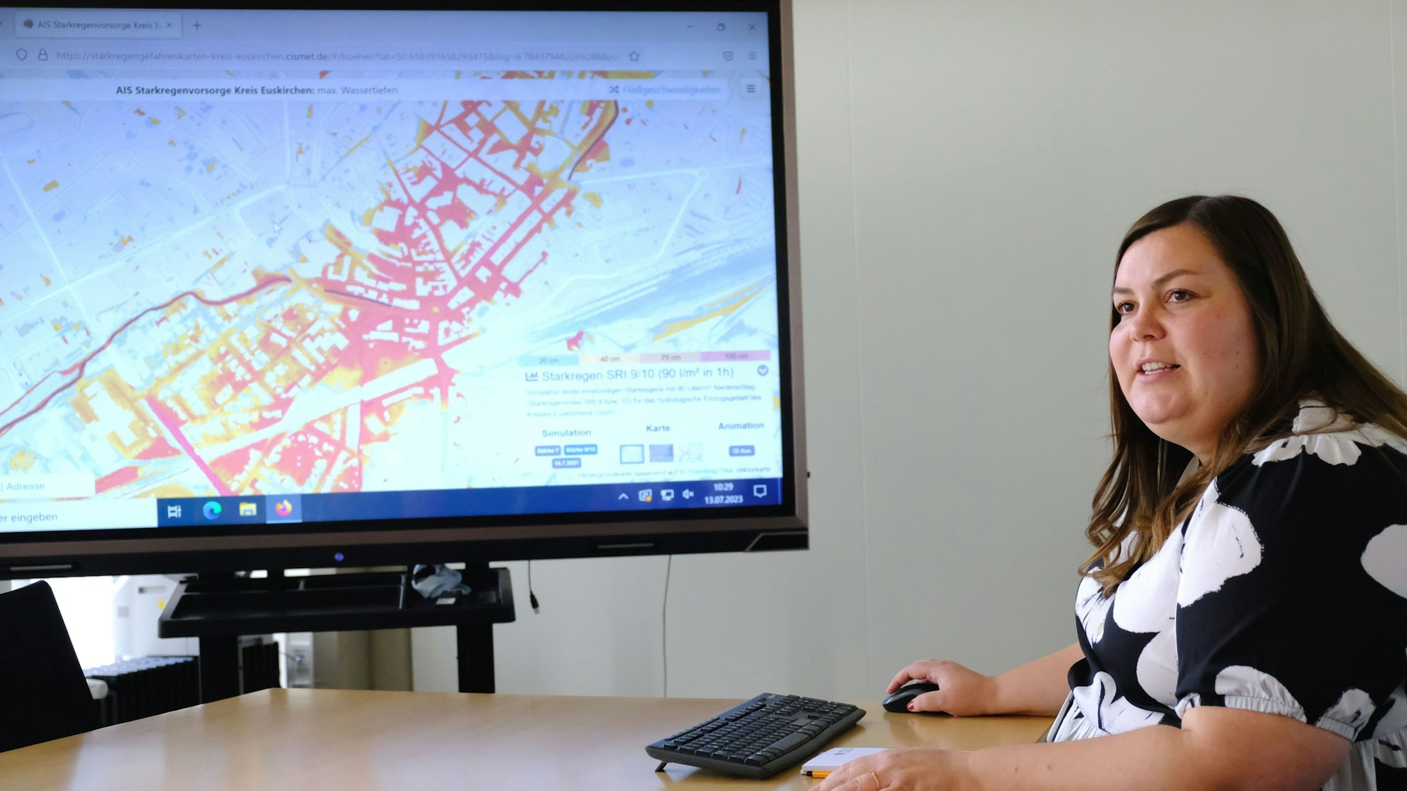 Eine Frau sitzt vor einem großen Bildschirm, auf dem eine Karte zu sehen ist. Sie trägt ein schwarz-weißes Oberteil und hat lange braune Haare.