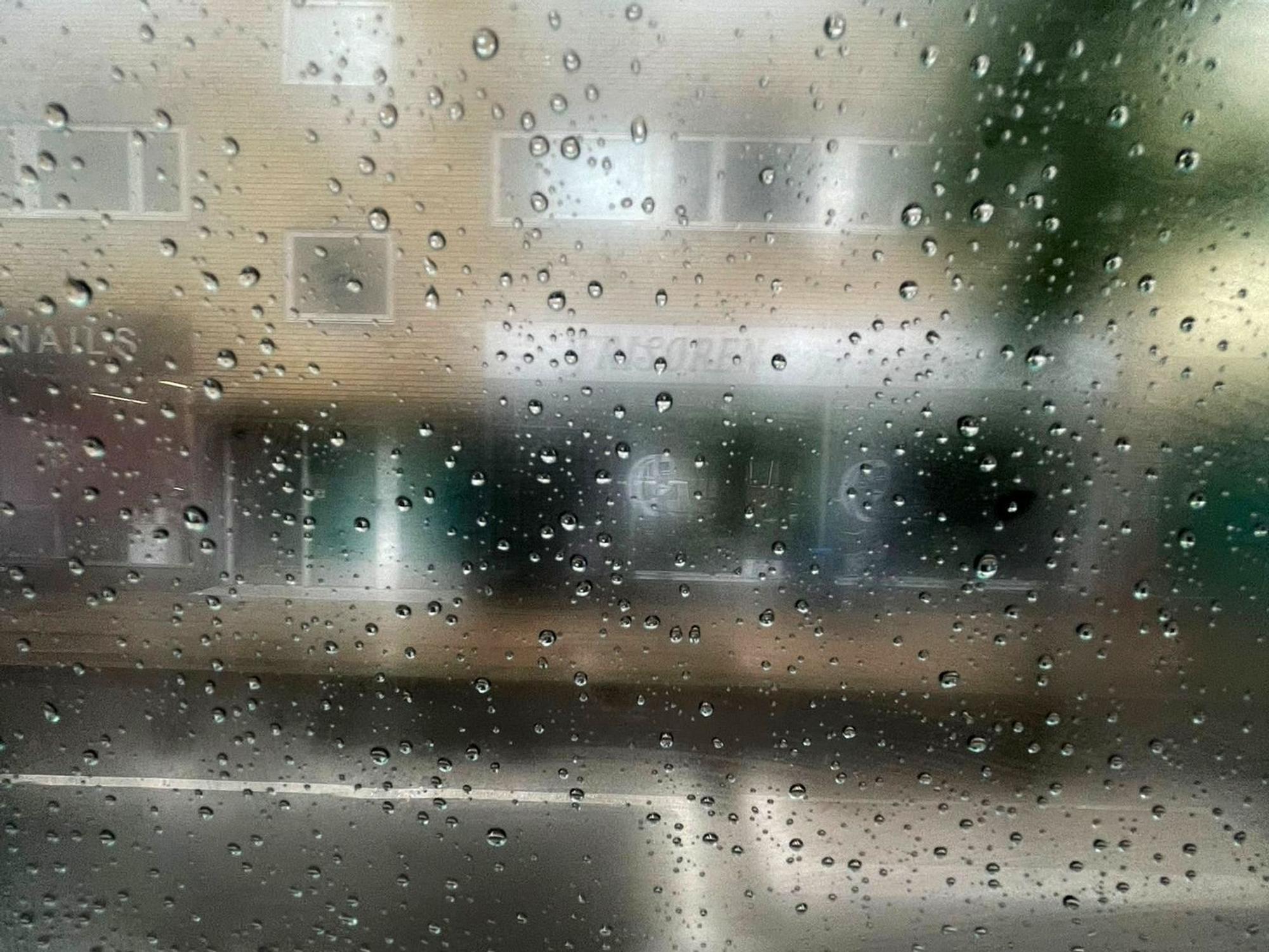 Regen auf einer Fensterscheibe, dahinter ist schemenhaft ein Gebäude zu erkennen.