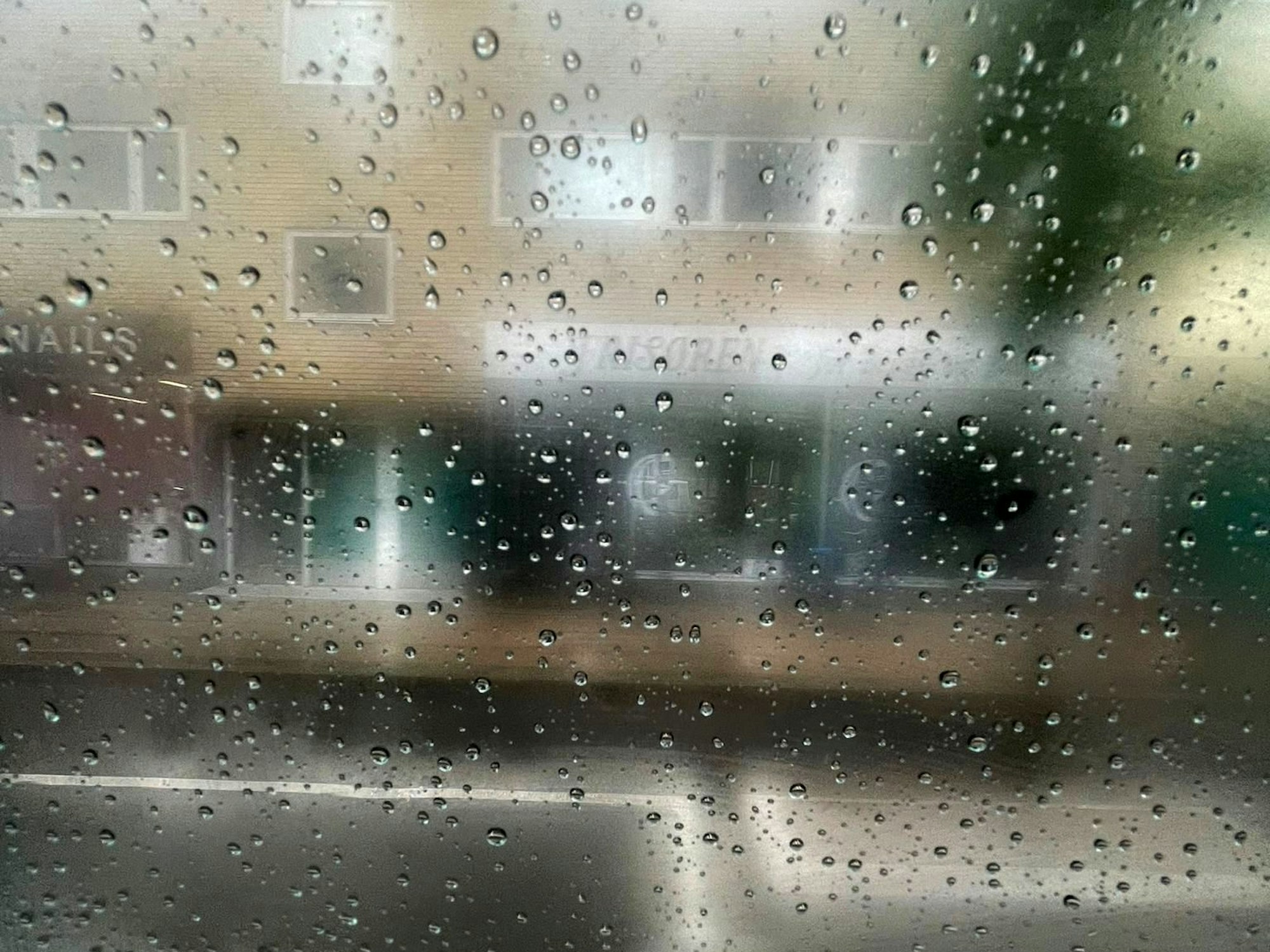 Regen auf einer Fensterscheibe, dahinter ist schemenhaft ein Gebäude zu erkennen.