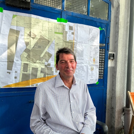 Ein Mann sitzt draußen. Hinter ihm ist ein Lageplan zu sehen.