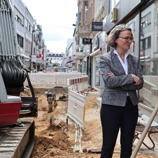 Die Euskirchener Innenstadt ist eine einzige Baustelle. Hier wird neues Pflaster verlegt. An der Baustelle, in der ein Kleinbagger steht, spricht Ina Scharrenbach mit der Praktikantin der Baufirma.