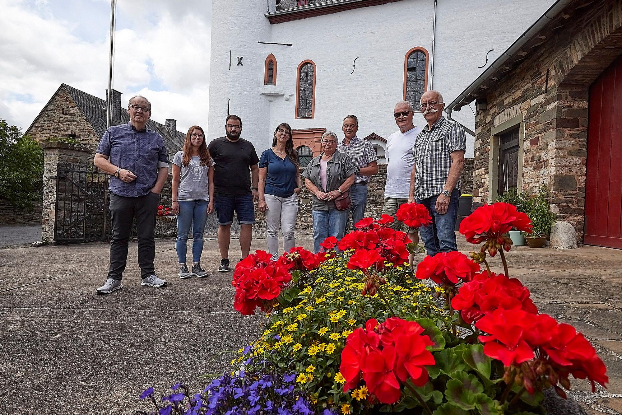 Eine Gruppe von Menschen steht auf einem Platz. Im Hintergrund ist ein Teil einer Kirche zu erkennen, im Vordergrund stehen gelbe, rote und lila Blumen.