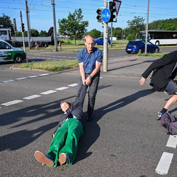 Ein Mitglied der Protestgruppe Letzte Generation wird bei einer Sitzblockade an der Autobahnabfahrt am Elbepark von einem Autofahrer (M) beiseite gezogen. Die Demonstranten tragen Masken mit den Portraits von Bundeswirtschaftsminister Habeck (l) und Bundeskanzler Scholz.