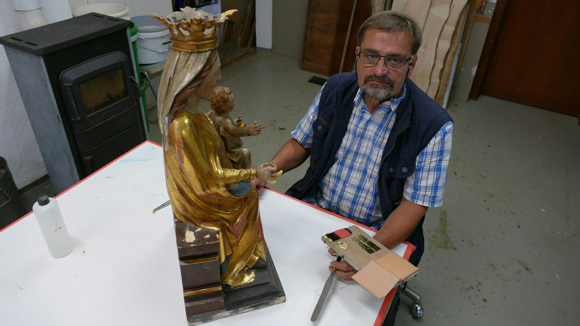 Restaurator Roland Gassert sitzt an seinem Arbeitstisch, vor ihm ist eine Marienstatue, an der er derzeit arbeitet.