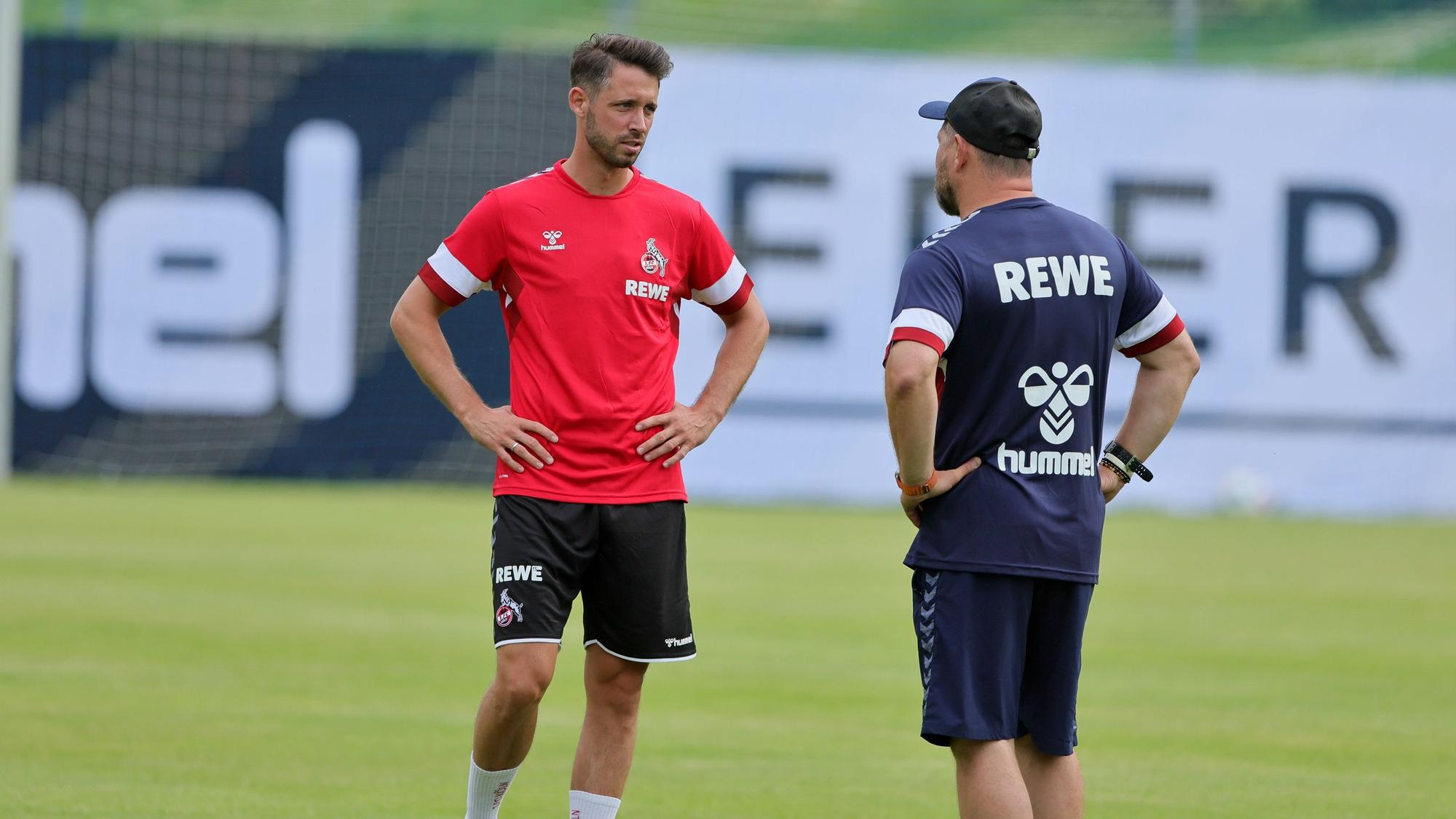 Mark Uth steht mit Steffen Baumgart auf dem Rasen und unterhält sich.