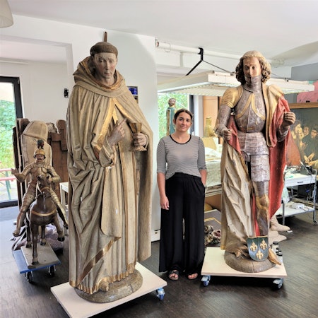 Eine Frau steht zwischen zwei großen Heiligenfiguren in einem Atelier.