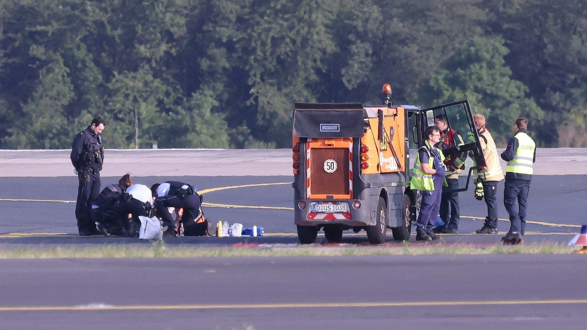 Sicherheitspersonal und Polizisten stehen auf dem Flugfeld und versuchen Aktivisten der Gruppe Letzte Generation vom Asphalt zu lösen, nachdem sie sich festgeklebt haben.