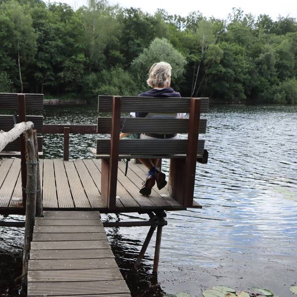 Eine Frau sitzt auf einer Bank, die auf einem Steg steht. Sie lässt den Blick über einen See schweifen.