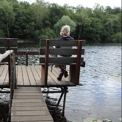 Eine Frau sitzt auf einer Bank, die auf einem Steg steht. Sie lässt den Blick über einen See schweifen.