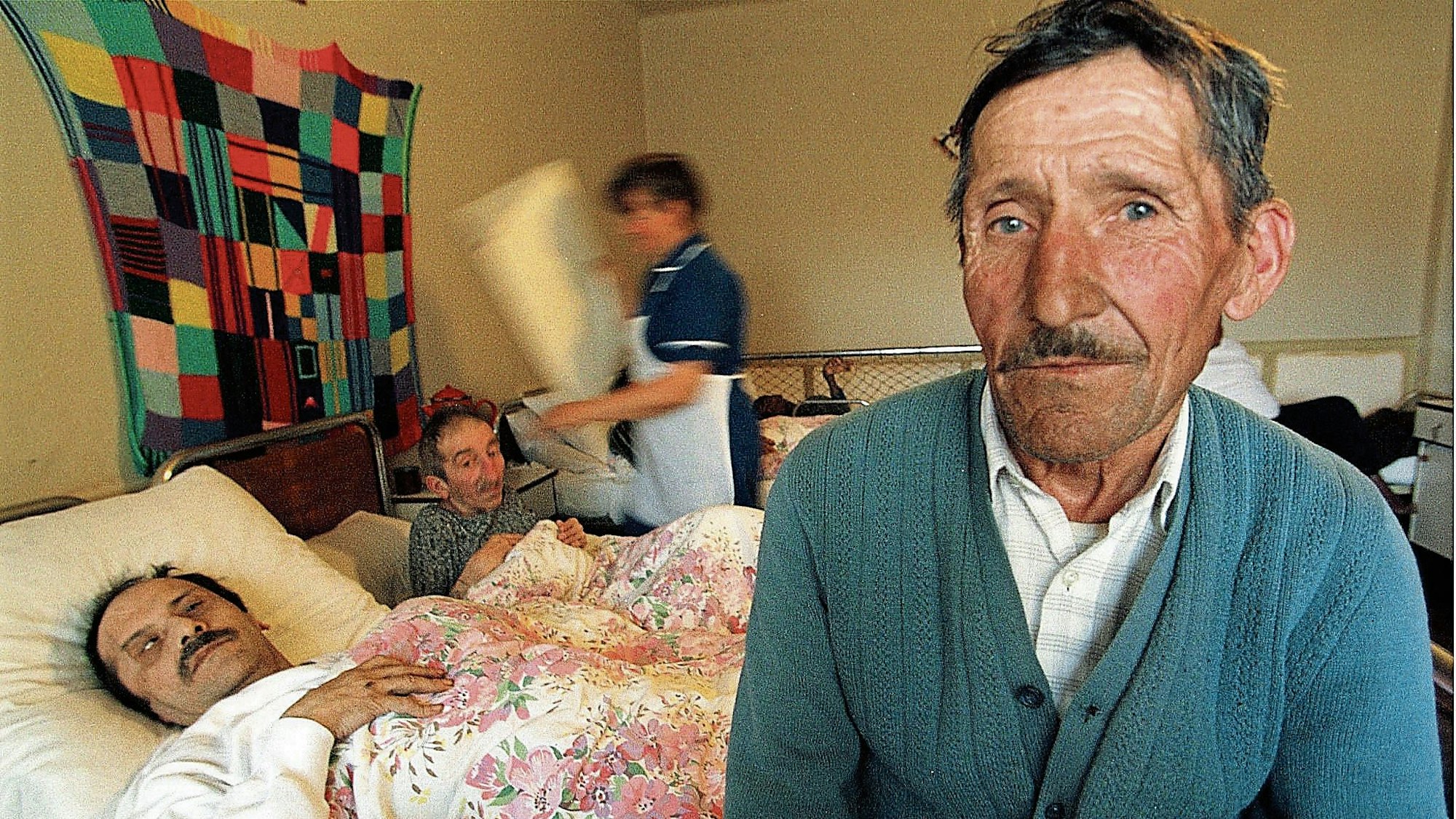 In die Kamera blickt ein dünner, alter Mann mit faltigem Gesicht. Hinter ihm sind zwei Männer zu sehen, die in einfachen Krankenhausbetten liegen.