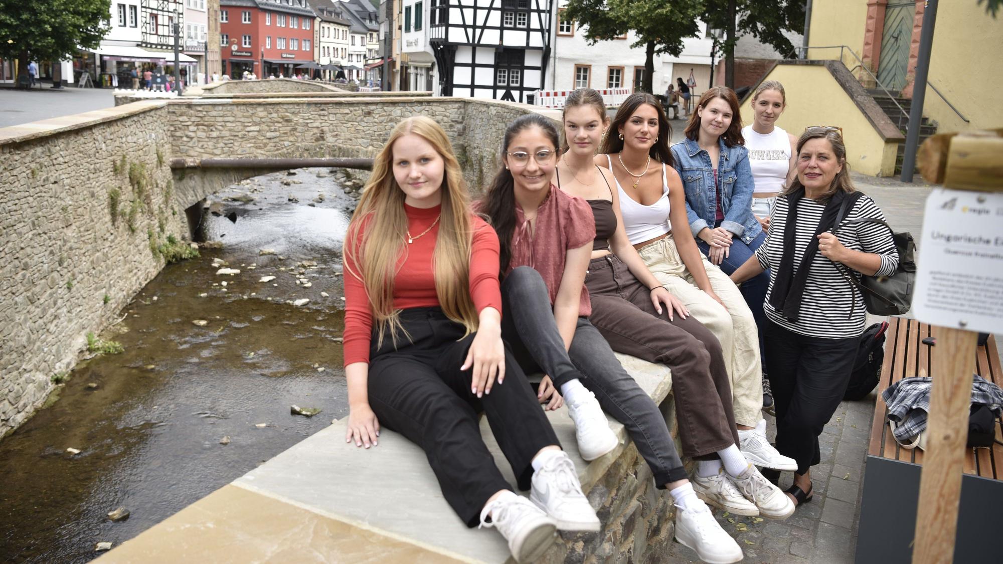 Sechs junge Frauen sitzen auf einer Mauer, neben ihnen steht eine Frau. Im Hintergrund sind ein Fluss und Fachwerkhäuser zu erkennen.