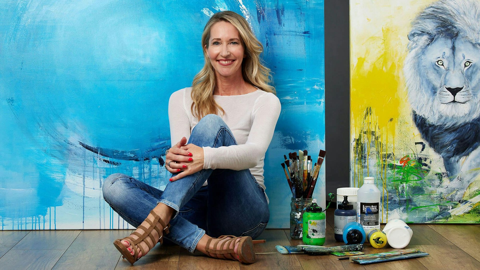 Die Künstlerin Susanne Herbold sitzt vor einer blau-gelben Leinwand, neben ihr stehen Farben und liegen Pinsel.