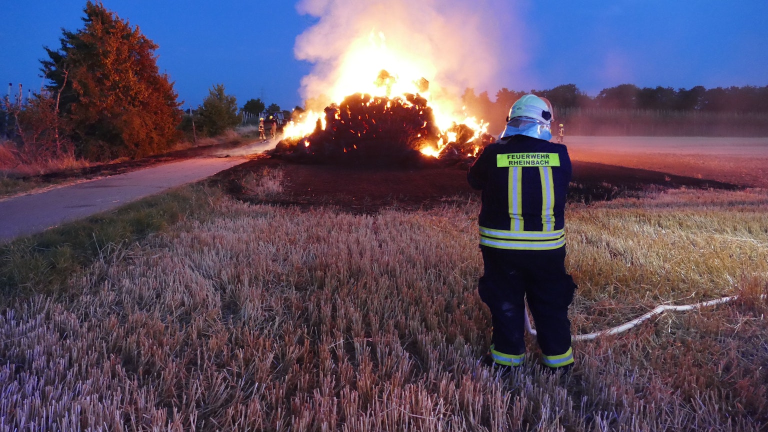 Im Hintergrund ist ein brennender Strohhaufen zu sehen, vorne steht ein Feuerwehrmann, der den Brand kontrolliert.