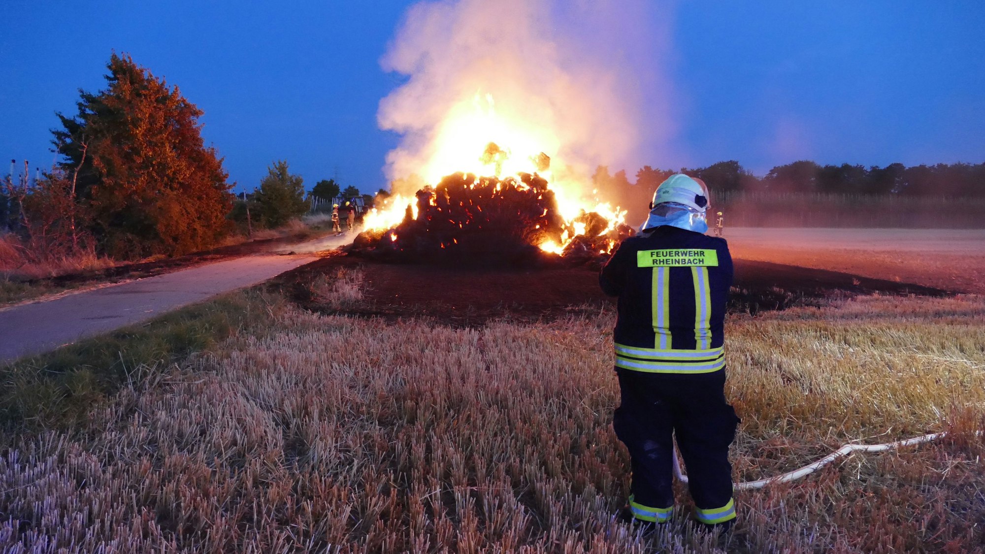 Im Hintergrund ist ein brennender Strohhaufen zu sehen, vorne steht ein Feuerwehrmann, der den Brand kontrolliert.