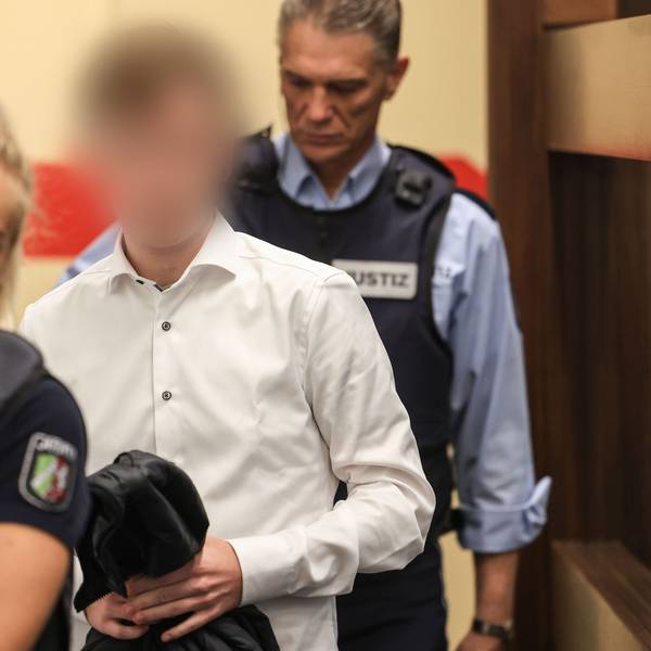 Der Angeklagte trägt ein weißes Hemd und wird von zwei Justizangestellten in den Gerichtssaal geführt.