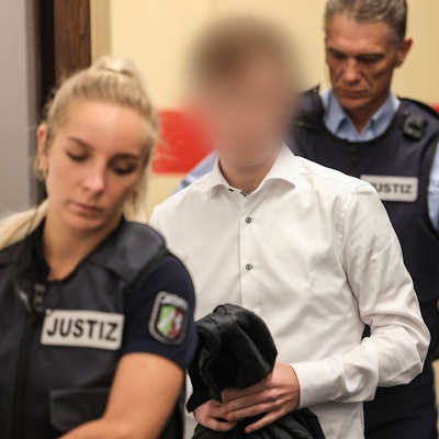 Der Angeklagte trägt ein weißes Hemd und wird von zwei Justizangestellten in den Gerichtssaal geführt.