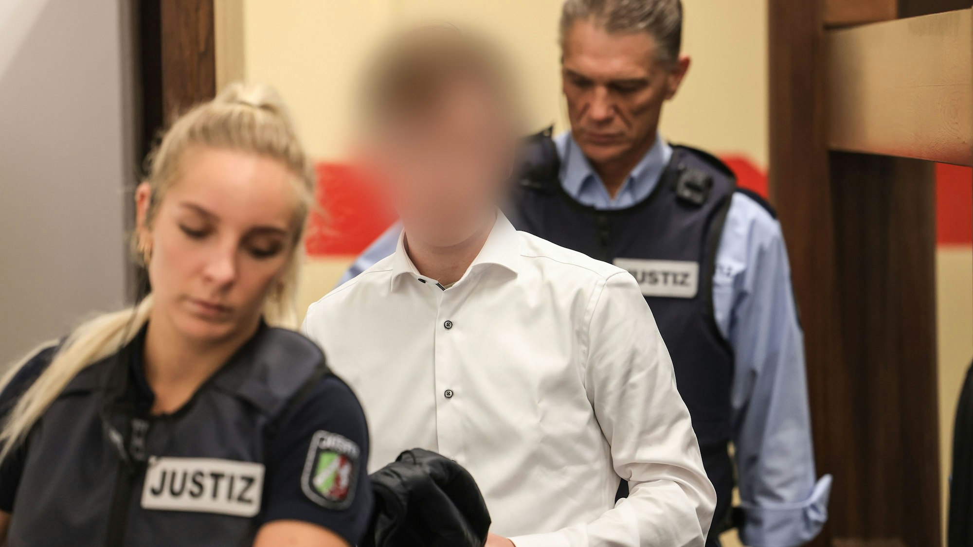 Der Angeklagte trägt ein weißes Hemd und wird von zwei Justizangestellten in den Gerichtssaal geführt.