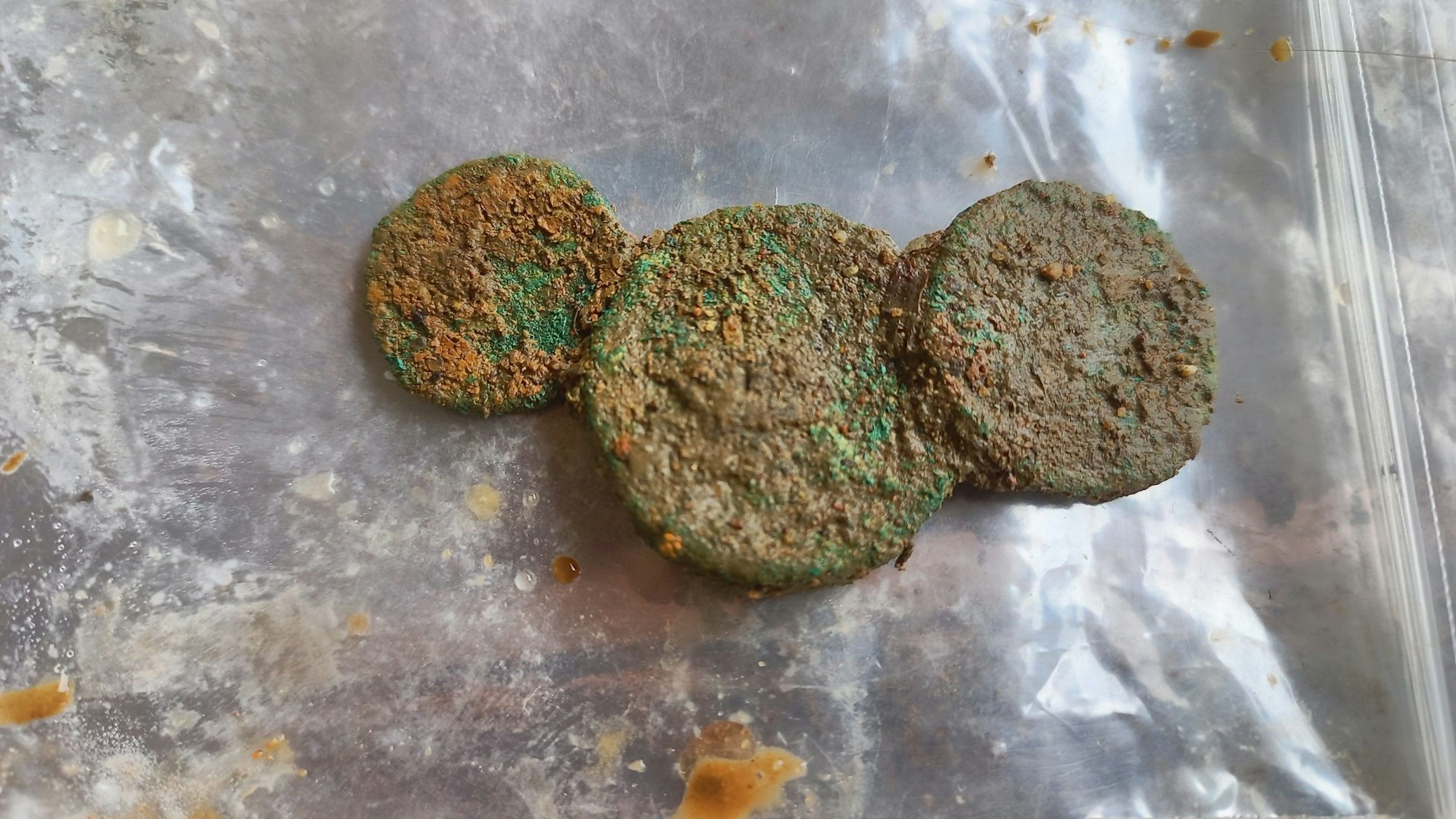 Die drei korrodierten Bronzemünzen auf einer Folie. Die Münzprägung ist nicht zu erkennen.