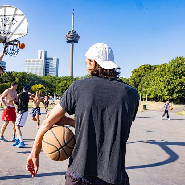 Mehrere Männer stehen auf einem Basketballplatz. Im Hintergrund ist der Colonius zu sehen.