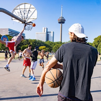 Mehrere Männer stehen auf einem Basketballplatz. Im Hintergrund ist der Colonius zu sehen.