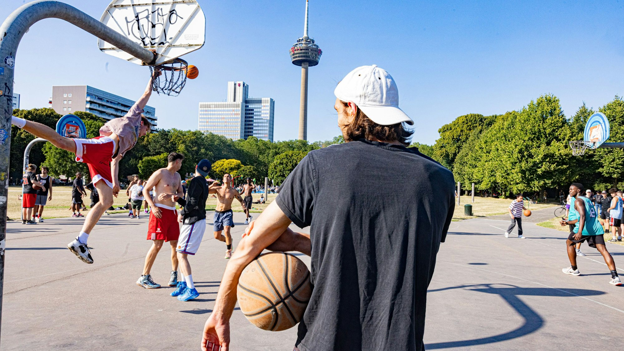 Mehrere Männer stehen auf einem Basketballplatz. Im Hintergrund ist der Colonius zu sehen.