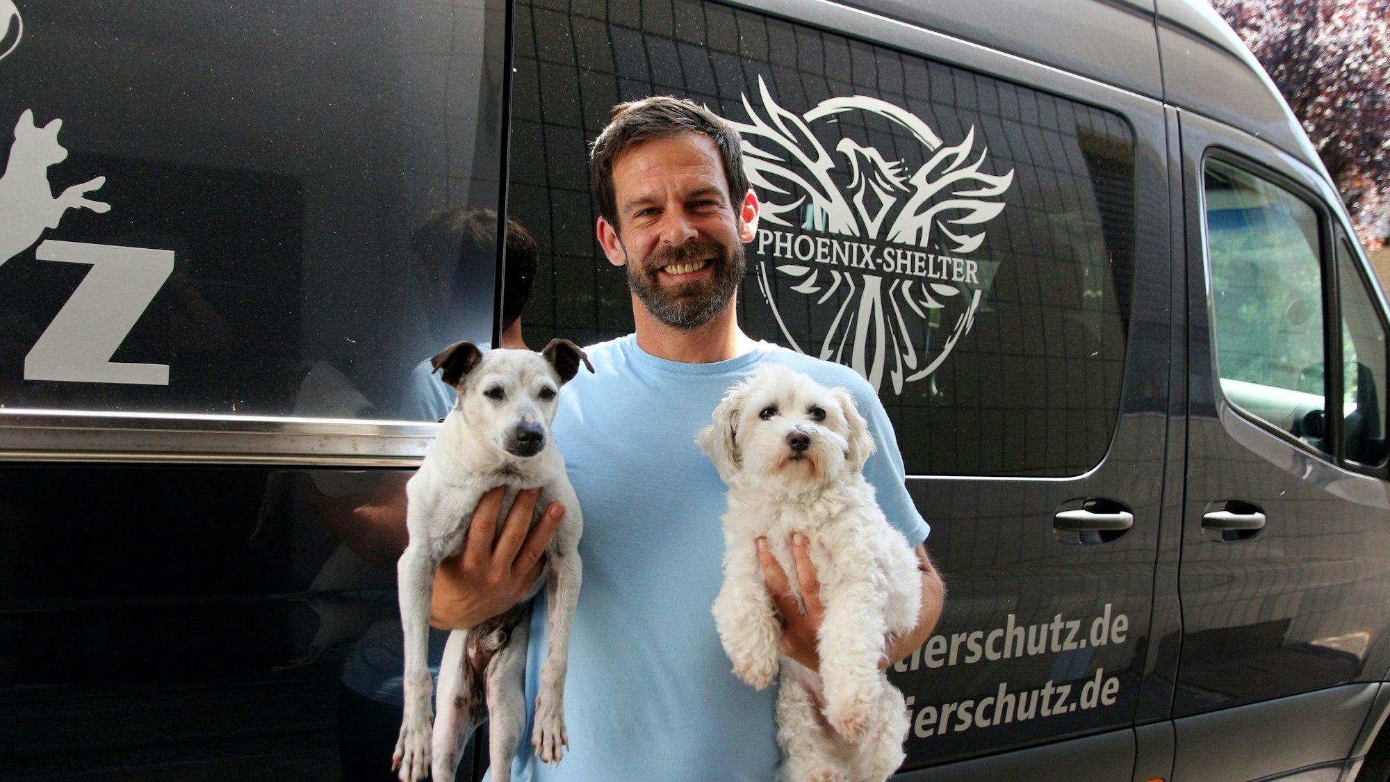Marcel Tong strahlt in die Kamera, in jedem Arm hält er einen Hund mit hellem Fell.