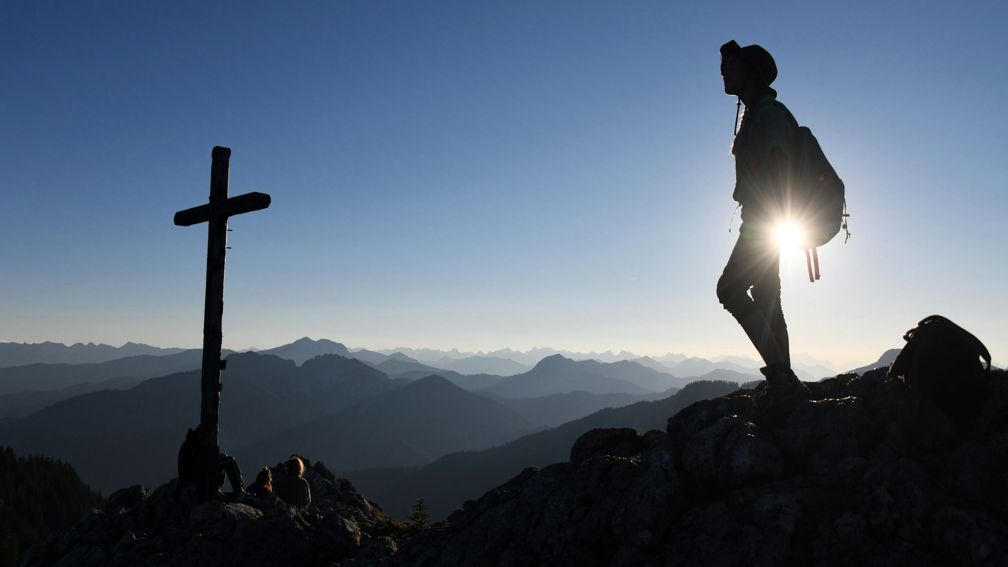Eine Ausflüglerin genießt den Blick, während am Gipfelkreuz des Taubensteins im Spitzinggebiet einige Wanderer pausieren.