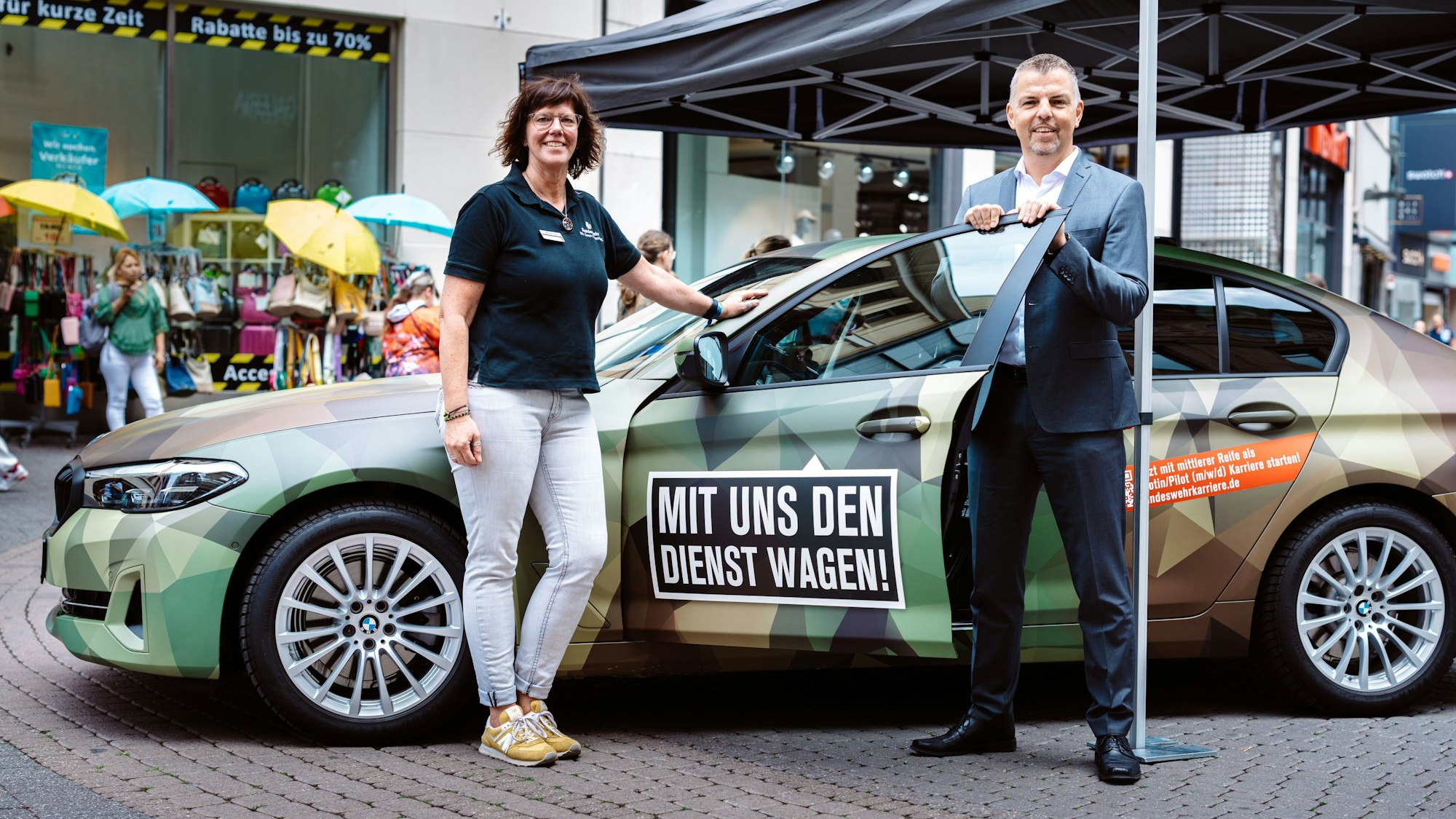 Nicole Venn und Thorsten Roth stehen vor einem in Tarnfarben folierten PKW auf der Schildergasse.