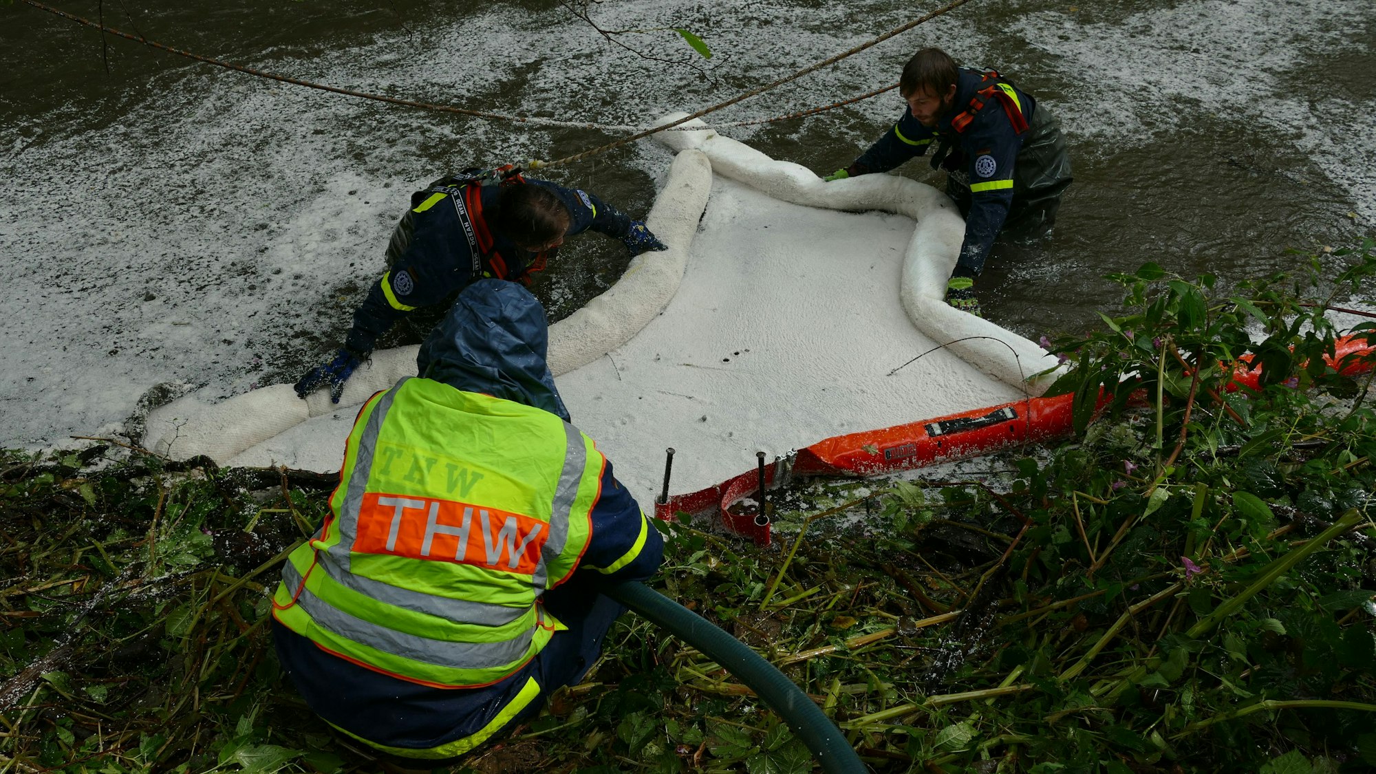 Im Oktober 2022 war das Öl im Wahnbach aufgefallen. Drei Einsatzkräfte des THW stehen bis zur Brust im schäumenden Wasser und errichten eine Ölsperre.