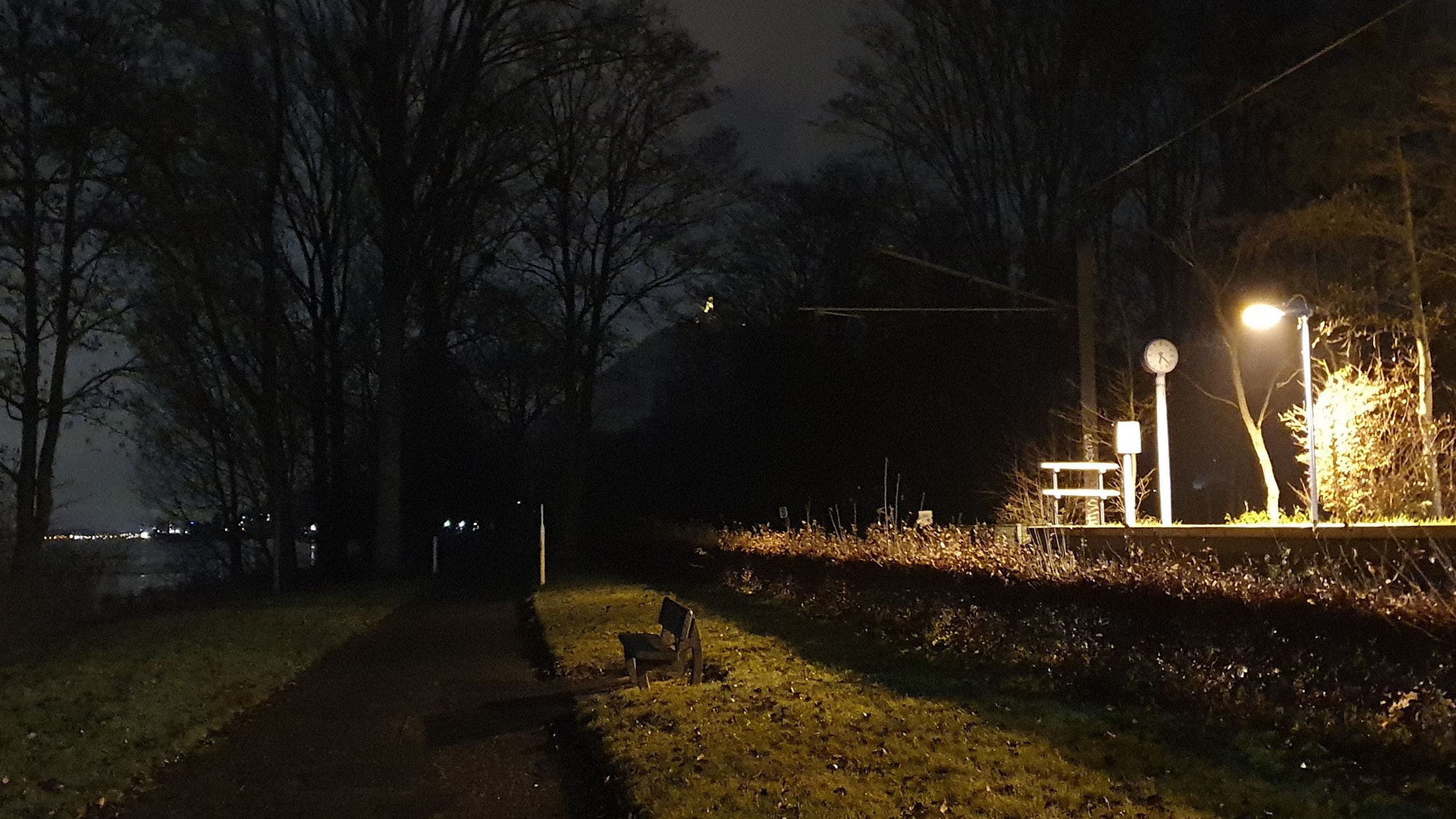 Ein Weg am Rheinufer führt in die Dunkelheit. Am rechten Rand kommt Licht von der Stadtbahnhaltestelle Am Splitzenbach.
