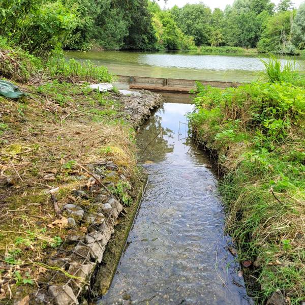 Das Foto zeigt einen schmalen Wasserlauf, die Überlaufschwelle, und im Hintergrund einen der Aufbereitungsteiche.