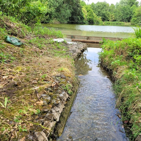 Das Foto zeigt einen schmalen Wasserlauf, die Überlaufschwelle, und im Hintergrund einen der Aufbereitungsteiche.