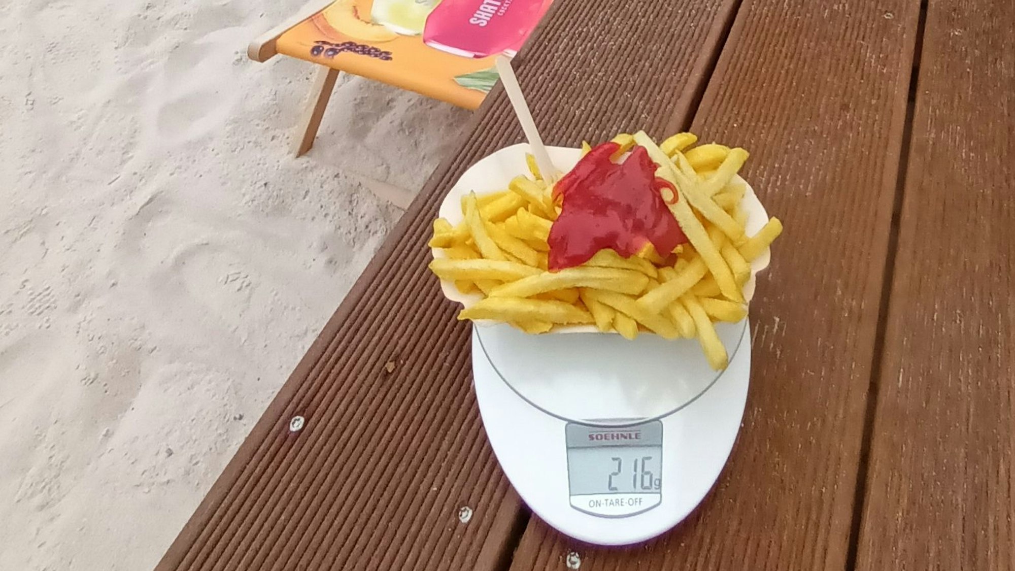 Pommes mit Ketchup stehen auf einer Küchenwaage, im Hintergrund ist ein Liegestuhl im Sand zu sehen. Die Waage zeigt 216 Gramm.
