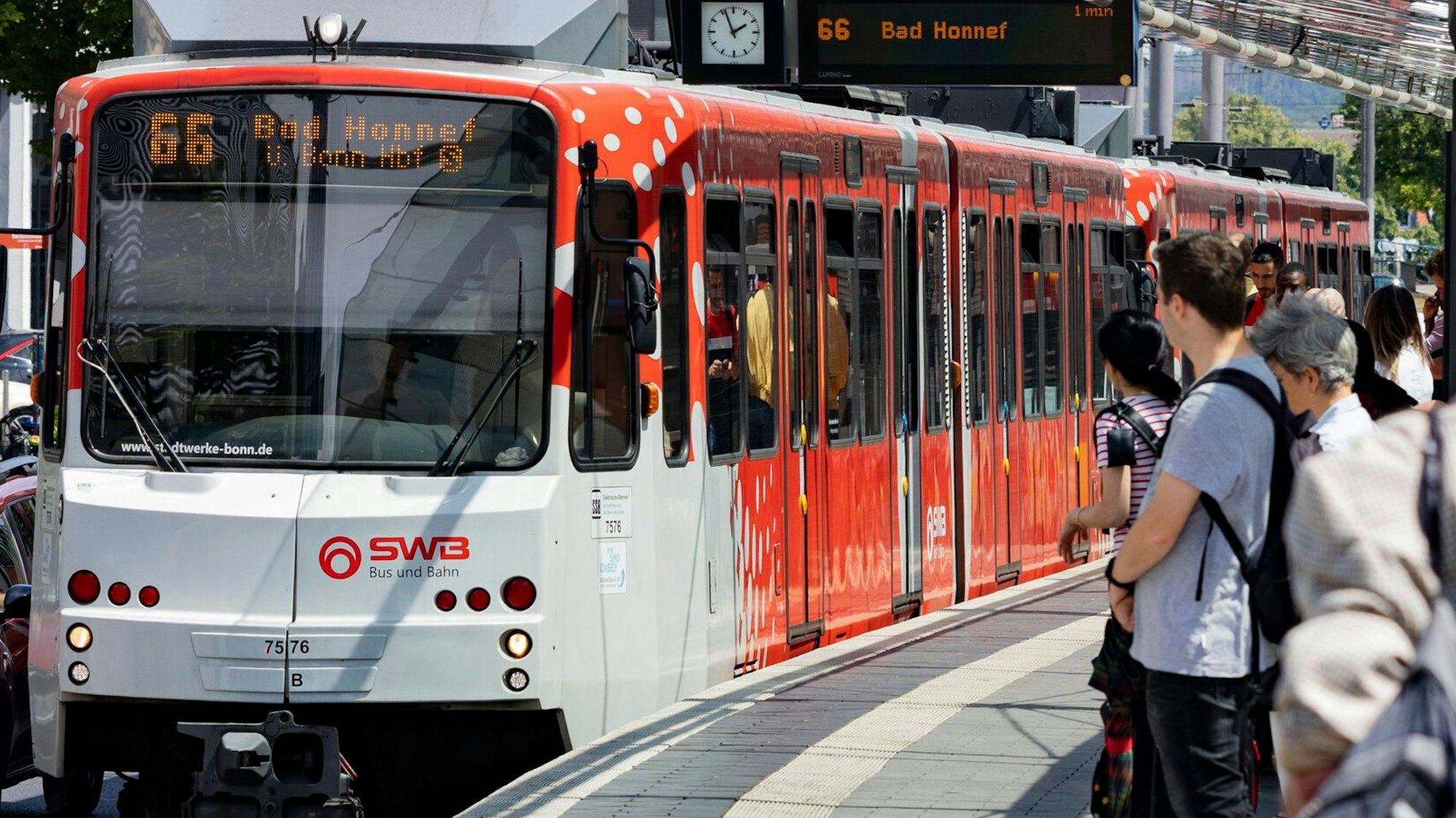 Eine rot-weiße Straßenbahn an einer Haltestelle. Menschen warten auf den Zug.