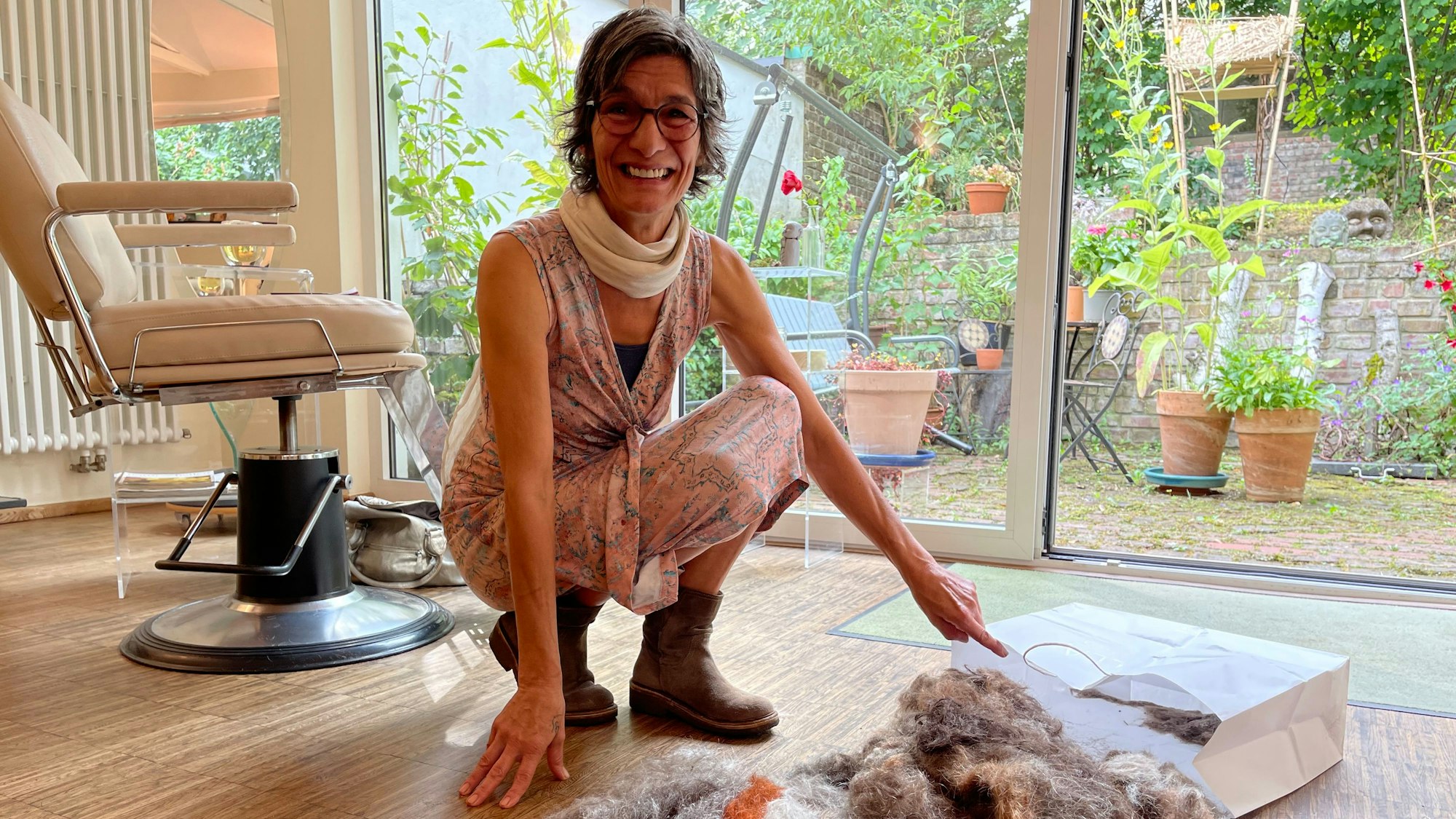 Eine Frau in einem Wickelkleid hat eine groß, geöffnete Papiertasche mit Haaren auf dem Boden ihres Salons gelegt.