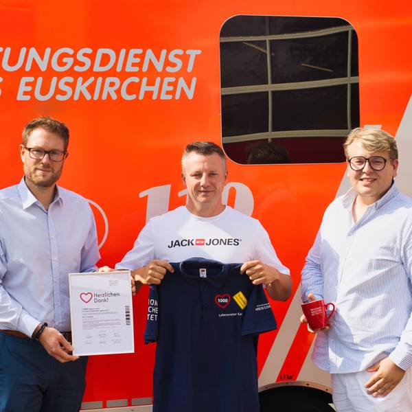 Drei Männer stehen vor einem Rettungswagen. Einer hält eine Urkunde in der Hand, einer ein T-Shirt, einer eine Tasse.