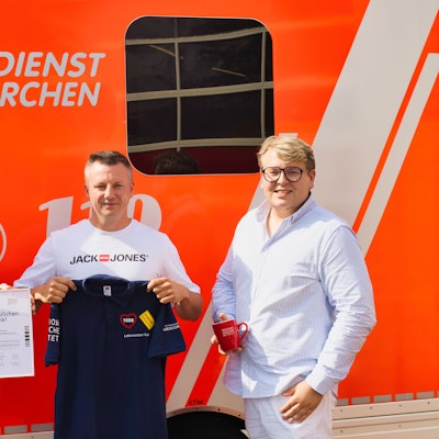 Drei Männer stehen vor einem Rettungswagen. Einer hält eine Urkunde in der Hand, einer ein T-Shirt, einer eine Tasse.